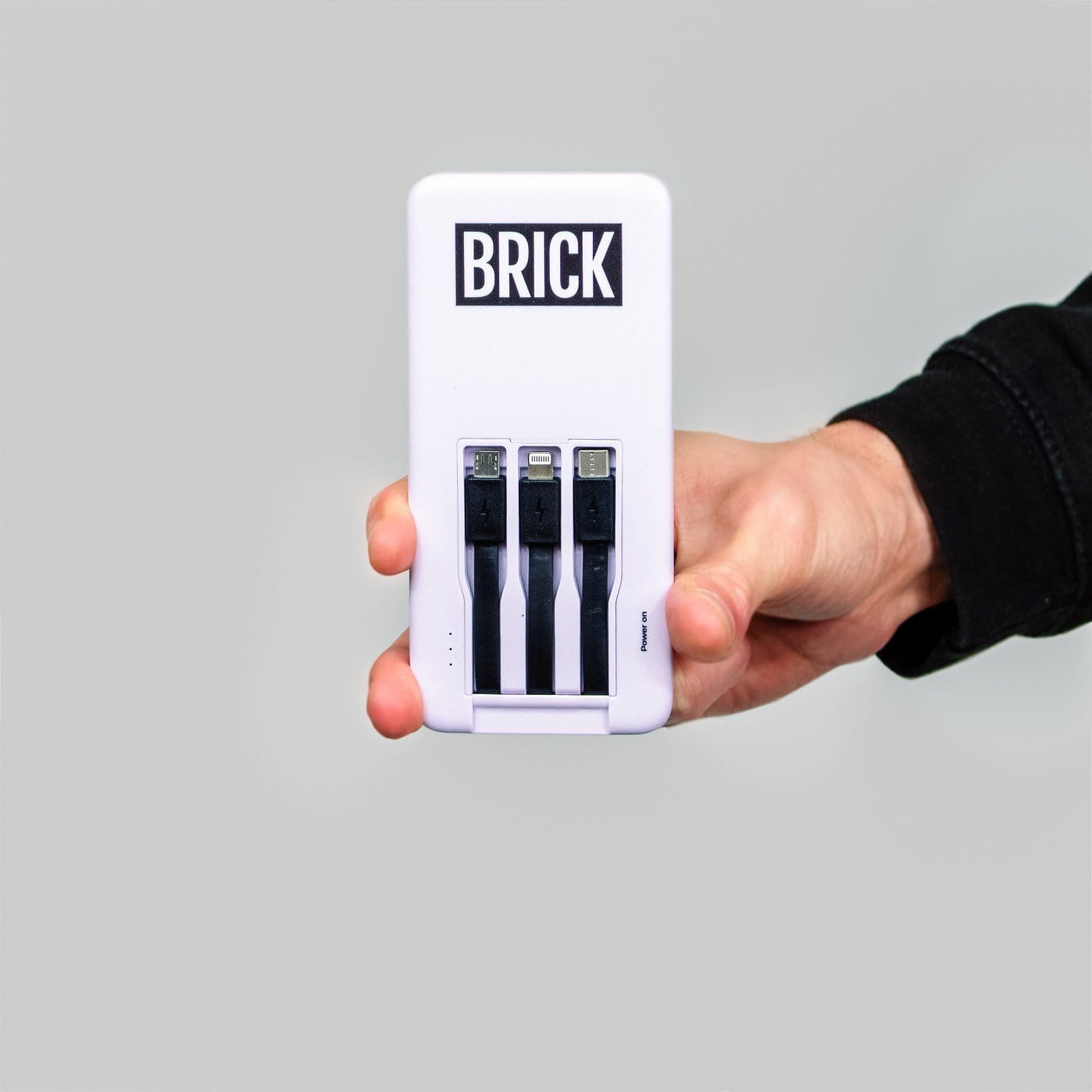 A hand holding a white Brick powerbank with its three built-in cables pulled out, on a light grey background